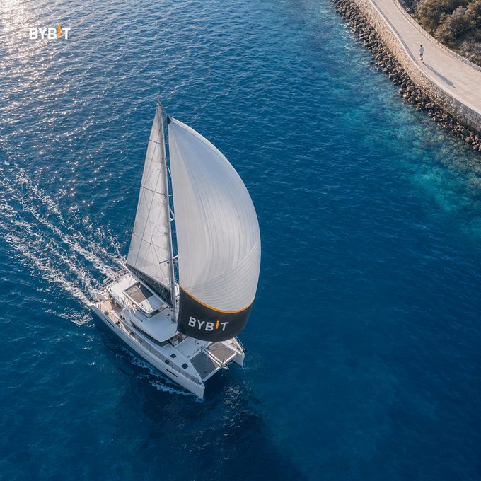 An aerial view of a white catamaran sailboat with a 'BYBIT' branded sail navigating blue waters near a rocky shoreline, with the Bybit logo also visible in the corner.
