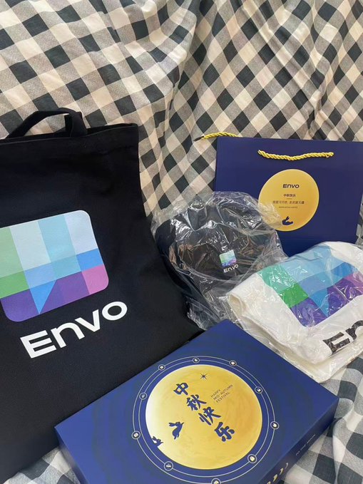 A collection of Envo branded merchandise on a checkered surface, including a black tote bag with a colorful triangle logo, a blue paper bag with a Mid-Autumn Festival moon design, and wrapped items including what appears to be a T-shirt.