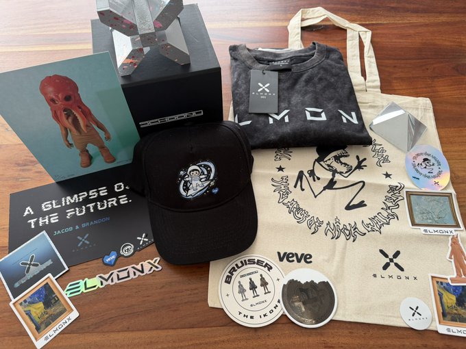 A collection of @ELMONX branded merchandise including a black cap, gray "ELMON" t-shirt, canvas tote bag, stickers, coasters, postcards featuring the "Jacob & Brandon" figure, and a branded box with a silver X sculpture, all arranged on a wooden surface. The swag haul showcases the collaboration between @elmonx_official and @veve_official, complementing the post celebrating Jacob Elmon's support following successful digital collectible drops like the Piet Mondrian x Doodles release and the VeVe Stickerverse platform launch.