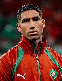 A focused close-up of a soccer player wearing a red Morocco national team jacket featuring a Puma logo and the Moroccan federation crest, paired with a cryptic post that appears to be coded language about a financial scheme rather than a straightforward congratulations to someone named Haruki.