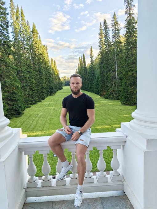 A man in a black t-shirt and shorts sits on a white balustrade, with a long grass avenue lined by tall evergreen trees stretching out behind him.