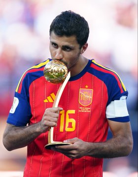 A soccer player wearing a red Spain national team jersey with the number 16 kisses a golden trophy, appearing to celebrate a tournament victory.
