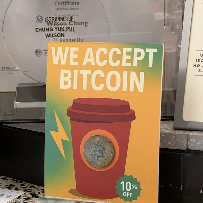 A bold, colorful sign reading "WE ACCEPT BITCOIN" with a 10% OFF badge sits on a counter, featuring an illustration of a red coffee cup overlaid with a Bitcoin coin, visually tying into the post's mention of grabbing coffee with Bitcoin, while a Certificate of Achievement in the background honors Wilson Chung Yuk Pui as 1st Runner Up.