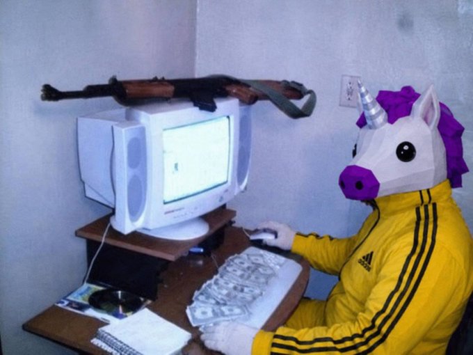 A person wearing a unicorn mask and a yellow Adidas jacket sits at a desk using an old CRT computer, with cash spread across the keyboard and an AK-47 resting on top of the monitor.