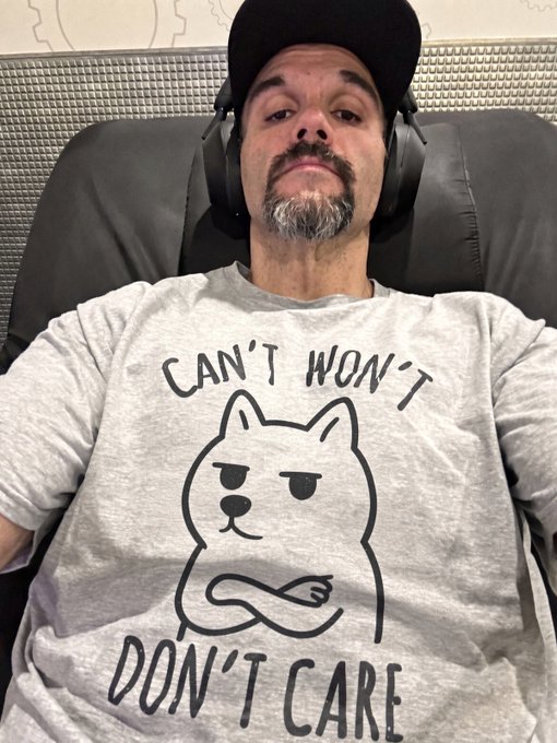 A selfie of a man wearing headphones and a black cap, sporting a gray t-shirt with a grumpy cat illustration that reads 'CAN'T WON'T DON'T CARE'.