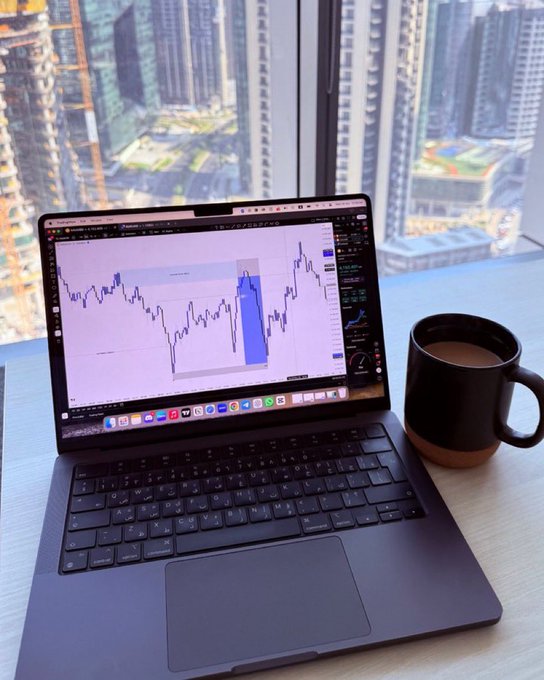 A laptop displaying a stock trading chart with a blue highlighted section sits next to a black mug of coffee on a desk overlooking a city skyline through a large window.