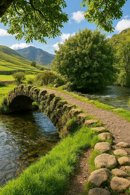 A scenic landscape featuring an old stone arch bridge over a clear stream, bordered by a dirt path lined with stones, vibrant green grass, lush trees, and rolling mountains in the background.