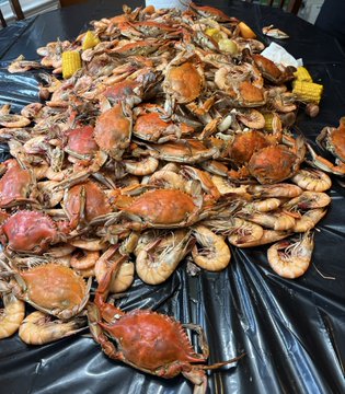 A large seafood feast spread across a black plastic-covered table, featuring a heaping pile of blue crabs, shrimp, corn on the cob, and potatoes.