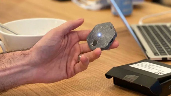 A hand holds a hexagonal gray Bitkey hardware wallet with a small LED light illuminated on its surface, shown on a wooden desk next to a coffee mug, laptop, and packaging box. The image illustrates the post's mention of the Bitkey device, highlighting its compact, stone-like design as part of the reviewer's positive first impression of the hardware-based Bitcoin self-custody solution using 2-of-3 multisig.