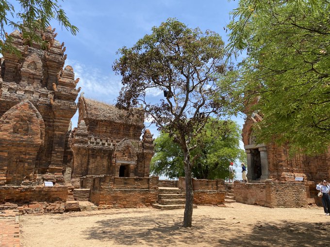 Ancient brick temple ruins, likely Khmer-style, stand under a bright blue sky with a tree in the courtyard and visitors visible near the structures.