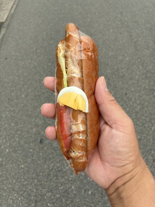 A hand holds a plastic-wrapped sandwich on a baguette containing egg, tomato, and lettuce against a pavement background.