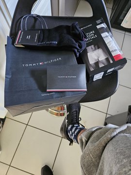 A pair of white Tommy Hilfiger high-top sneakers on a black box with shopping bag, photographed on a tiled floor with a person's feet visible.