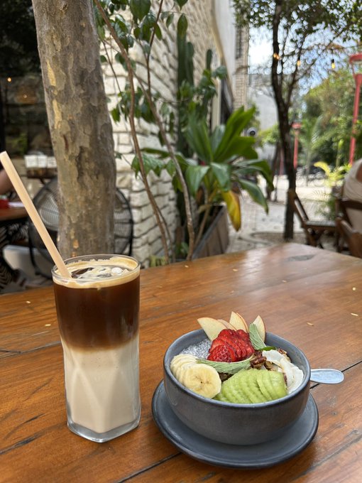 An iced coffee drink with a bamboo straw next to a bowl of acai or smoothie topped with fresh strawberry, banana, and apple slices, served on a wooden table at an outdoor cafe.