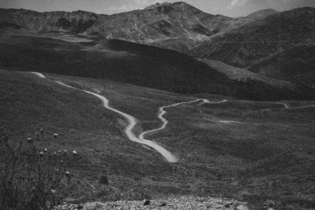 A black and white photograph of a winding dirt road cutting through barren hills with mountains visible in the distance.