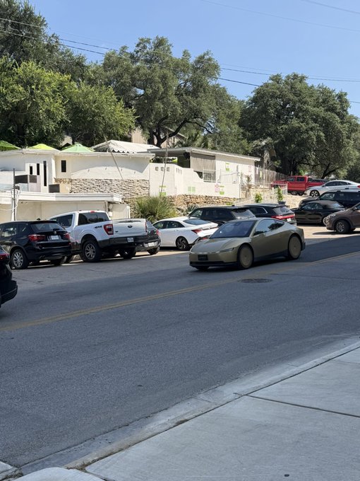 A street scene showing parked vehicles including a gold Nissan 370Z, with houses and trees visible in the background.