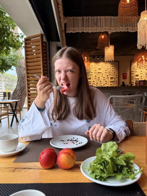 A woman with long brown hair wearing a white embroidered blouse makes a strained facial expression while eating something red with a fork at a restaurant table; the table holds a plate of greens, two red fruits, a small dish with seeds, and a cup of coffee.