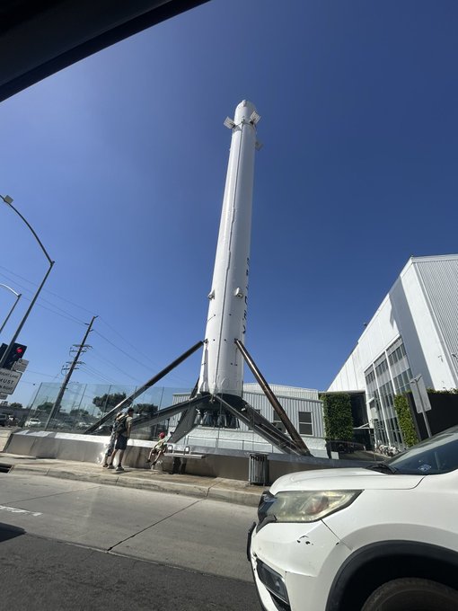 A large white SpaceX Falcon 9 rocket booster stands vertically on display outdoors, mounted on black landing legs with a person standing nearby for scale and a white car in the foreground.