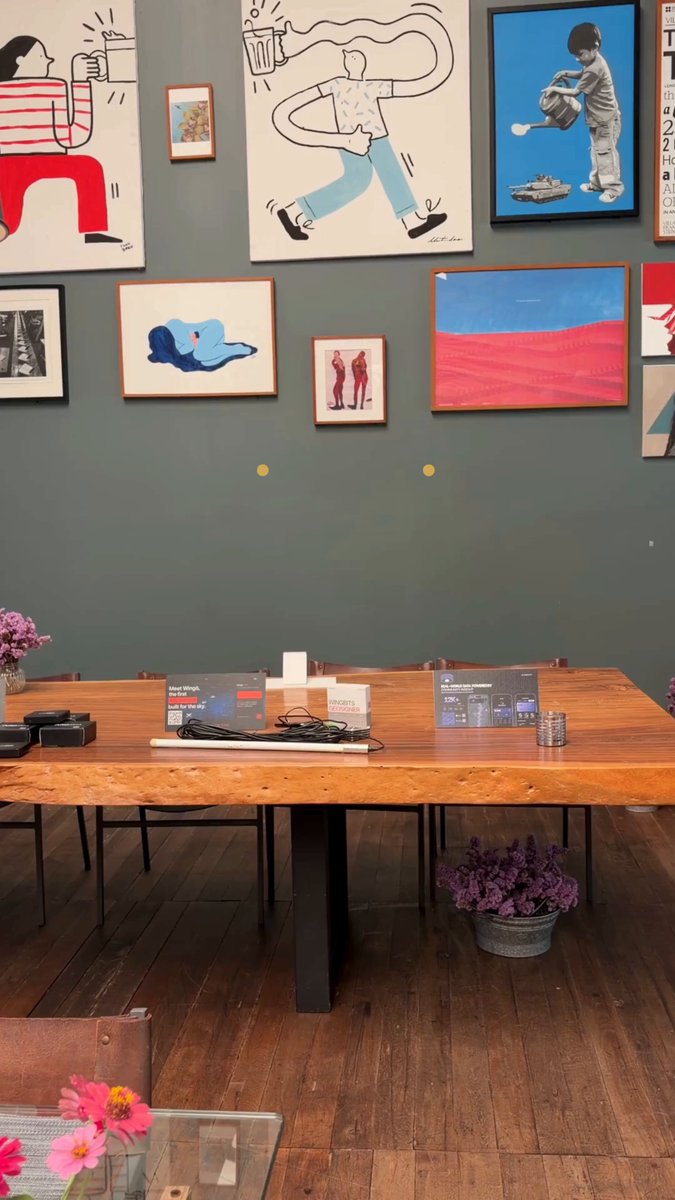 The image features a large wooden dining table with chairs surrounding it, set against a gray wall adorned with various paintings. The room also contains potted plants placed around the space, adding a touch of greenery to the scene. A person can be seen in the background, possibly admiring or preparing for the meal that will take place at this inviting and artistic dining area.