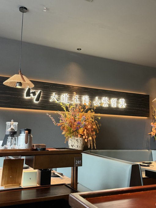 Interior of a Japanese restaurant featuring a backlit wooden sign with kanji characters, a hanging woven pendant lamp, and a dried floral arrangement on a dark wall above booth seating.
