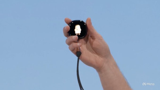 A hand holds up a small, circular black device with a glowing central component and an attached cable, set against a clear blue sky background. The "Meta" logo appears in the bottom right corner, visually aligning with the post's claim about Meta launching a new palm-sized 5G AI device.
