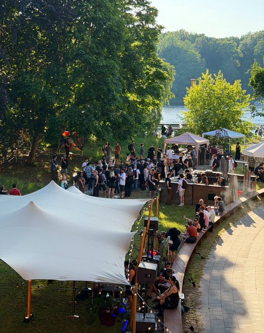 An outdoor daytime event beside a lake, featuring a crowd of people gathered near a stage area under large white tent canopies and string lights, with lush green trees in the background.