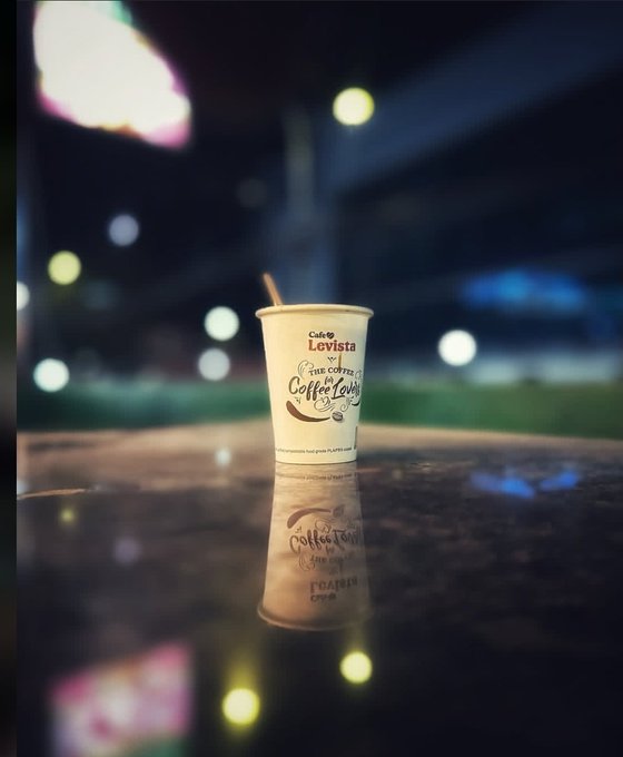A nighttime photograph of a Levista coffee cup with a straw sitting on a dark reflective table, with blurred city lights in the background creating a reflection.