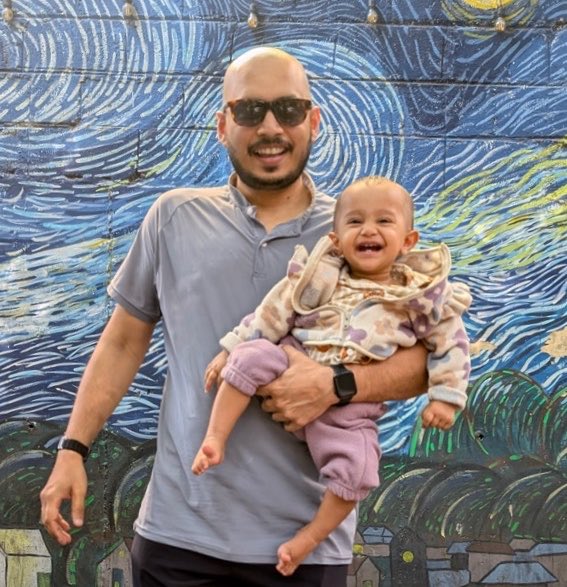 A smiling bald man with sunglasses holds a laughing baby in a patterned hoodie in front of a colorful mural resembling Van Gogh's Starry Night.