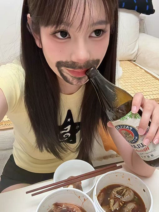 A selfie of a young woman wearing a yellow shirt, holding up a beer bottle to her face in a playful pose, with bowls of food and chopsticks in front of her.