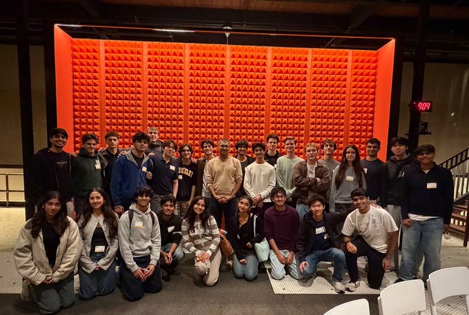 A large group poses in front of an orange, pyramid-textured acoustic wall in an indoor venue.