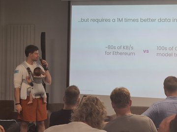 A man wearing a baby carrier stands on a stage giving a presentation, with a slide behind him mentioning 'Ethereum' and '1M times better data.'