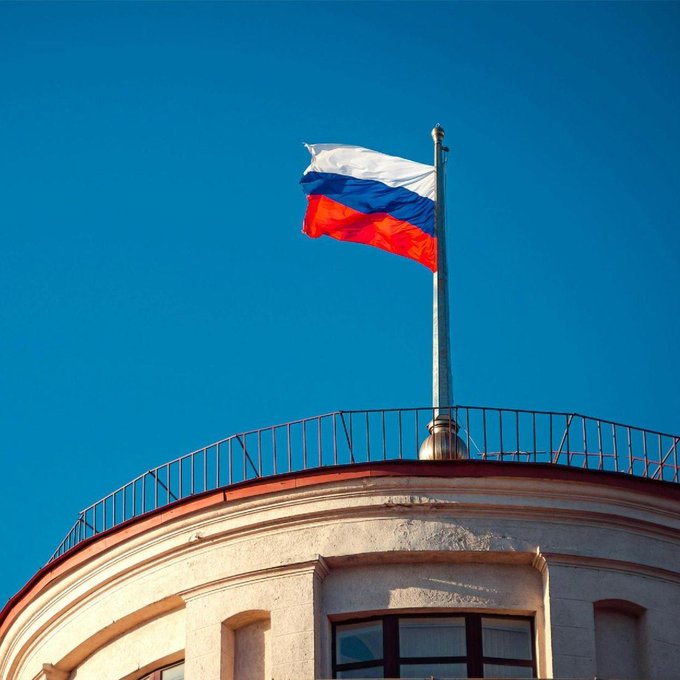 The Russian flag (white, blue, and red horizontal stripes) waves on a flagpole atop a circular building against a clear blue sky.