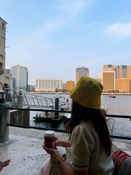A view from behind of a person wearing a yellow bucket hat holding a coffee cup, looking out over a waterfront with a city skyline and boats in the distance.