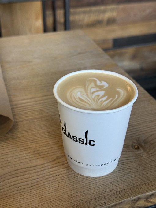 A white paper cup containing a latte with leaf-patterned latte art sits on a wooden table; the cup features the word 'CLASSIC' with a flame logo.