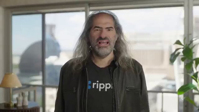 The image features a man with long hair wearing a black jacket standing in front of a window. He has a serious expression on his face as he looks directly at the camera. The background shows an office setting, which includes a desk and a potted plant. There is also a lamp visible in the scene. The post that accompanies this image reads: "JUST IN: Ripple CTO Joel Katz says enterprises are already using $XRP Ledger for real-world asset tokenization".