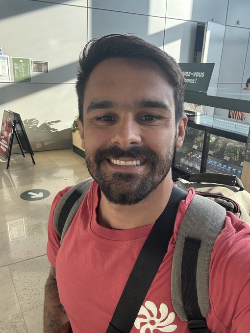 Selfie of a smiling bearded man wearing a red t-shirt and backpack, taken inside what appears to be an airport terminal.