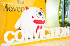 A white mascot character with a red smile stands next to a large "CONVICTION" sign at the TRON booth, with a yellow banner behind reading "Real" and "Moven," highlighting the exclusive merchandise pickup opportunity for community members engaging with the TRON ecosystem.