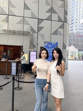Two women pose for a photo indoors, one in a cream top and jeans giving a thumbs up, the other in a white dress waving, with a modern building visible in the background.