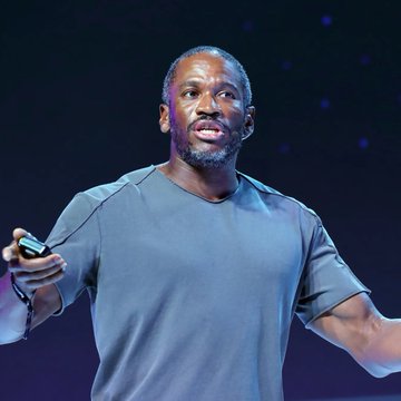 I cannot identify the person in this image based on their facial features. I can see a man with a beard wearing a gray t-shirt, appearing to be on stage giving a presentation, holding what looks like a remote or clicker. Without additional context indicating who he is, I cannot confirm whether this is Arthur Hayes mentioned in the post, so I won't write a caption making that identification.