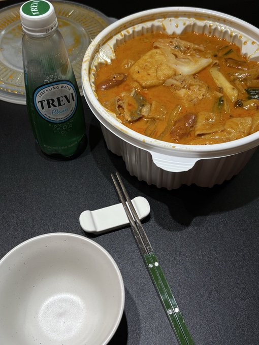 A takeout container filled with a spicy stew or soup containing tofu, vegetables, and meat, accompanied by a bottle of 'TREVI' sparkling water, chopsticks, and an empty bowl on a dark table.