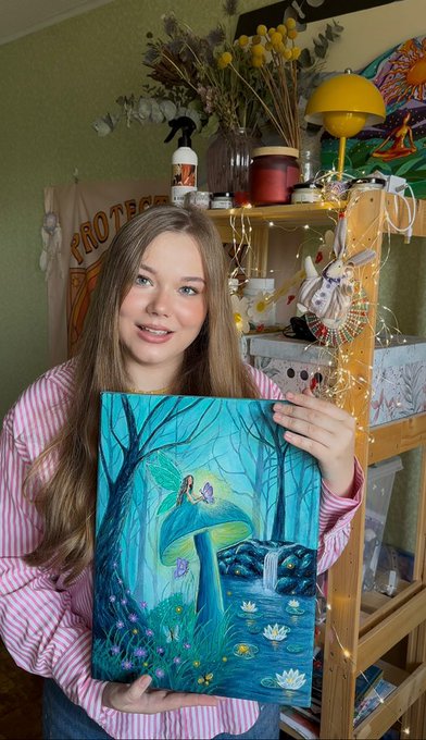 A young woman with long brown hair, wearing a pink striped shirt, holds up a vibrant teal-colored original painting featuring butterflies, a glowing mushroom, a waterfall, and lotus flowers in a mystical forest scene, which visually brings to life her poetic message about butterflies "woven from miracles" choosing you. The artwork's enchanting atmosphere and ethereal details directly illustrate the dreamlike sentiment shared in the post about life preparing something magical and unimaginable.