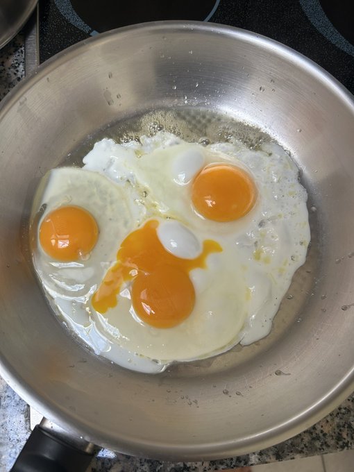 The image features three fried eggs cooking in a silver frying pan, placed on top of an oven. The eggs are cooked to perfection with their yolks runny and whites fully set. The background appears to be a kitchen countertop or stovetop, providing context for where the cooking is taking place. The caption "GM! This is what a 69 IQ face looks like. Keep on stackin' them sats".