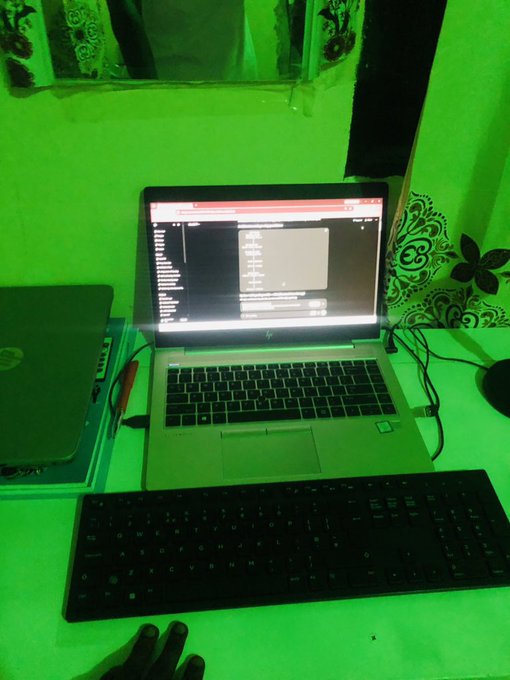A workspace with green ambient lighting featuring an HP laptop displaying code, an external black keyboard, and a closed laptop on the desk.