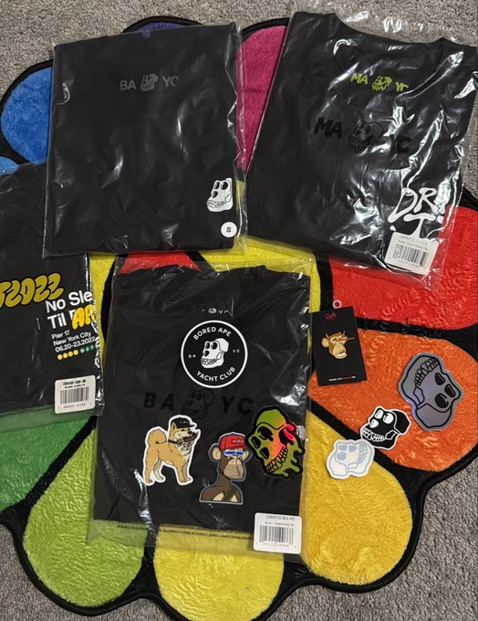Black 'BABY YC' and 'Bored Ape Yacht Club' t-shirts and accessories with monkey-themed stickers spread across a colorful Twister game mat.