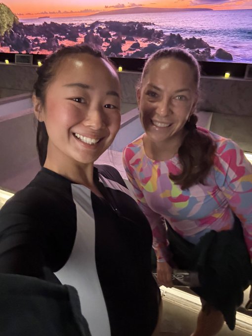 A selfie of two smiling women posing together, with a rocky coastline and sunset visible in the background.
