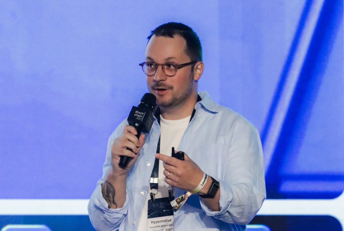 A man with glasses and short hair wearing a light blue shirt, holding a microphone and speaking on stage at what appears to be a Binance event, wearing a 'Hypematter' lanyard.