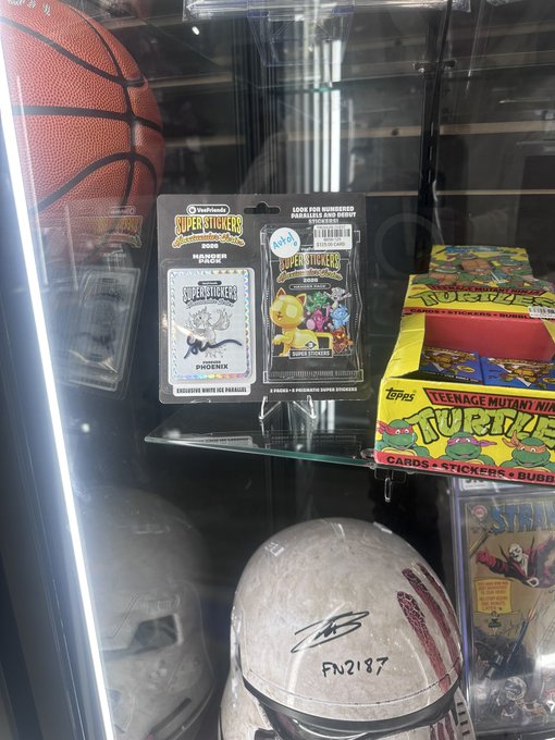 A glass display case containing collectibles including a basketball, Teenage Mutant Ninja Turtles cards and stickers, signed football helmets, and Superstickerz packets featuring Spider-Man, Phoenix, and Lion King characters.