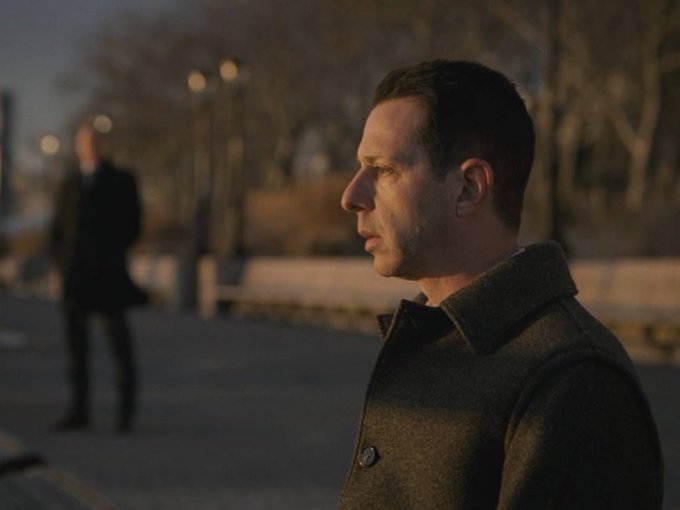A man in a dark coat stares blankly into the distance during golden hour, while another figure stands in the blurred background—visually capturing the somber, contemplative mood of Bitcoin investors reflecting on a downturn. The image evokes the quiet resignation and disbelief implied by the post's reference to Michael Saylor's controversial Bitcoin strategy.