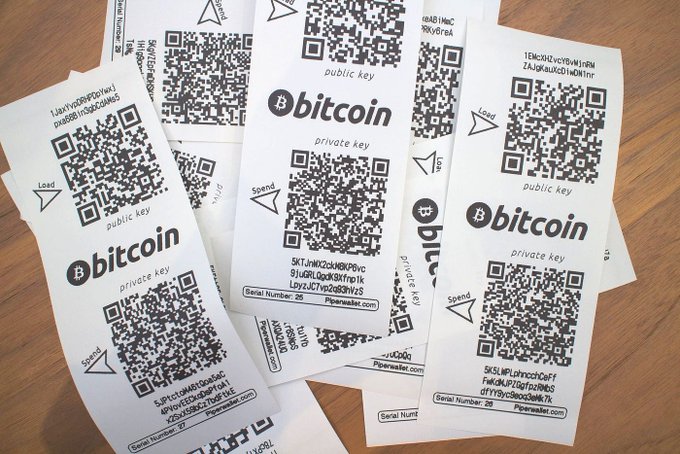 A scattered collection of paper wallets labeled "bitcoin" featuring QR codes for both public and private keys, with "Spend" and "Load" indicators, serial numbers, and the website "Paperwallet.com" visible.