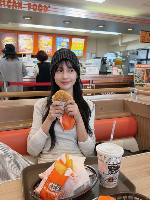 A young woman wearing a gray beanie and long-sleeve shirt sits in an A&W restaurant booth holding a burger, with fries and a root beer in a cup styled like a newspaper on the tray in front of her.