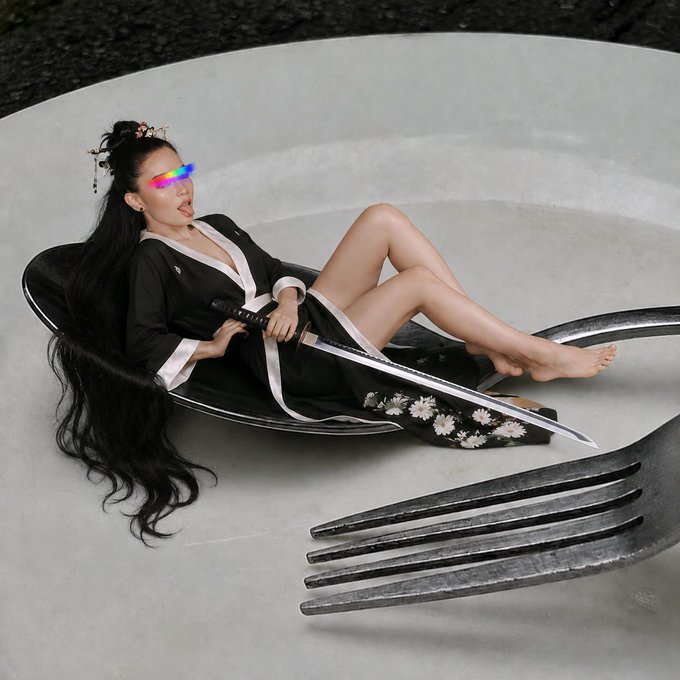 An artistic photo featuring a woman in a black and white robe reclining on a large spoon while holding a katana, with an oversized fork positioned below on a plate.